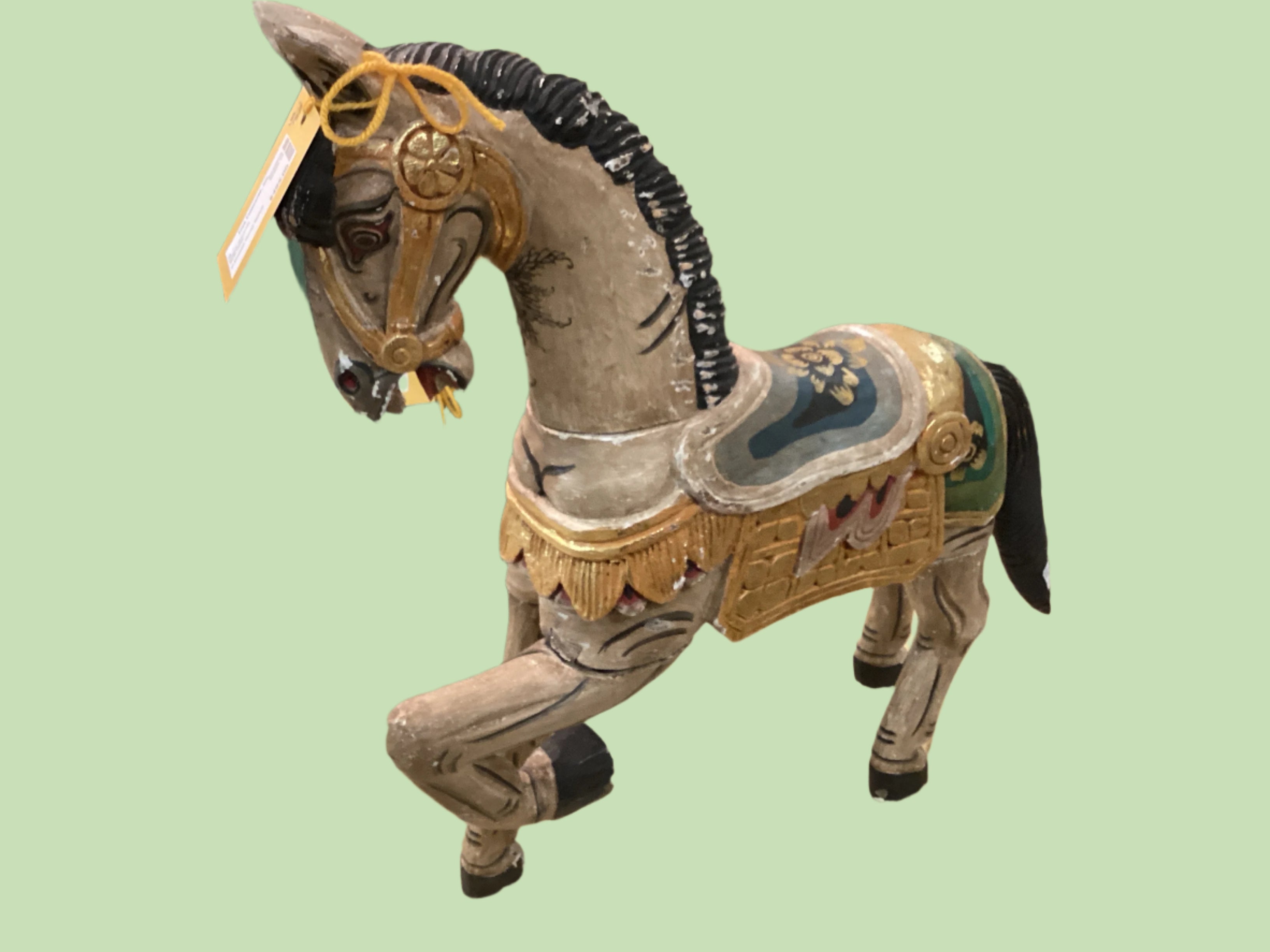 A multicolored vintage horse statue with decorative elements, likely made of wood and other materials, displayed against a backdrop of furniture and decorative items.